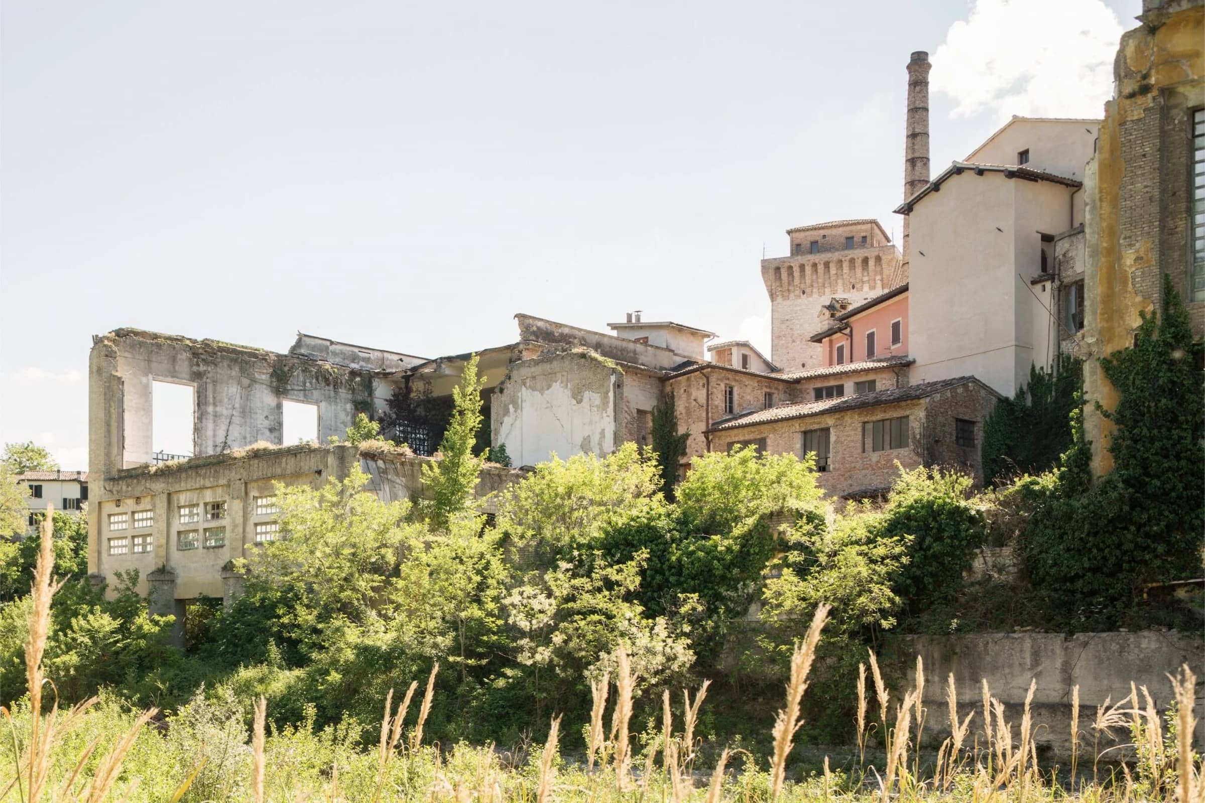 Abandoned industrial buildings with overgrown vegetation in the foreground, a smoking chimney in the background, and a partly cloudy sky above.